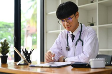 Beyaz üniformalı pratisyen hekimin portresi modern işyerinde oturuyor ve tıbbi form dolduruyor...