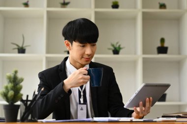 Kahve içen ve dijital tablet kullanan erkek ekonomist, ofis masasında satış istatistikleri belgesi ile çalışıyor..