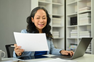 İstatistik belgeleriyle çalışan ve ofis masasında dizüstü bilgisayar kullanan başarılı bir orta yaşlı iş kadını...