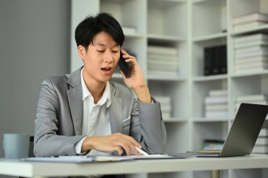 Asyalı erkek yatırımcı cep telefonuyla konuşuyor, iş ortağı modern işyeriyle pazarlık ediyor...