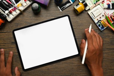 Sanatçının ahşap masada dijital tablet, akrilik boya tüpleri ve paletleri kullanışının üst görüntüsü..