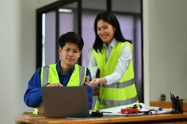 Yansıtıcı yelek giyen iki mühendis planlar üzerinde çalışıyor ve yeni projeyi ofiste tartışıyorlar..