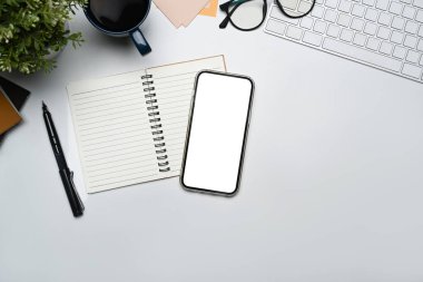 Beyaz ekranlı akıllı telefon, defter, kahve fincanı ve beyaz masadaki bardaklar. Üst manzara, düz uzanma.