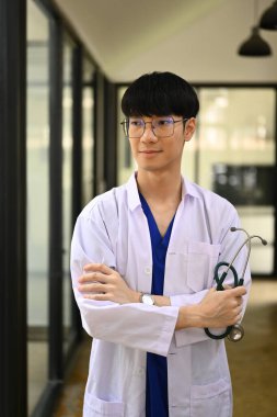 Pencerenin yanında duran beyaz önlüklü, erkek pratisyen hekimin portresi.