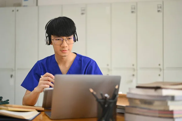 亚洲男性医科学生戴着耳机在笔记本电脑上观看在线讲座 医疗实习概念 — 图库照片