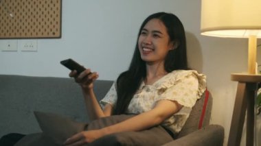 Ağır çekimde gülümseyen, sıradan kıyafetler giyen, kanepede dinlenen ve televizyon izleyen genç bir kadın..