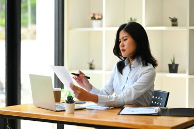 Ciddi Asyalı kadın ekonomist dizüstü bilgisayar kullanıyor ve iş yerinde istatistik raporu hazırlıyor