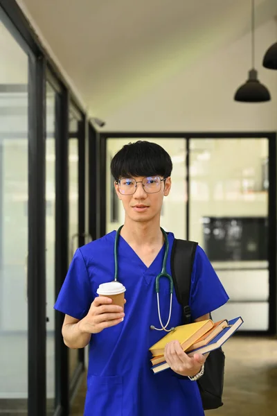 Portrait of asian male medical student in blue scrubs holding paper cup and looking at camera..