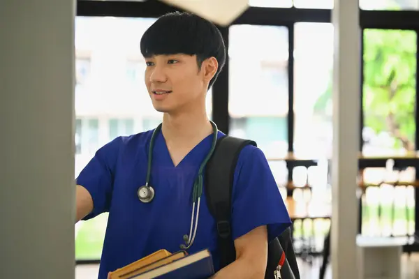 Medical student man wearing blue scrubs with backpack standing at lockers in campus.