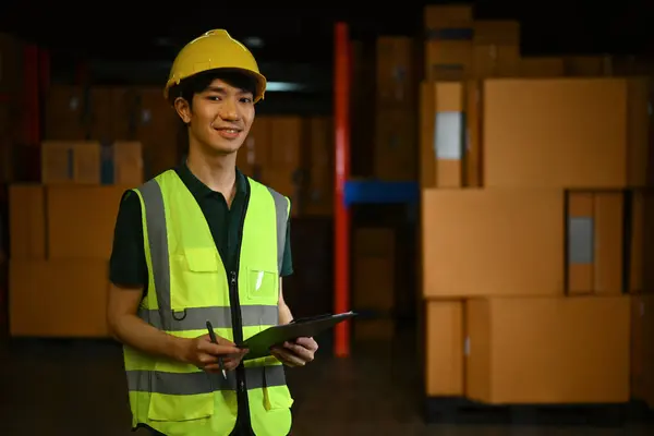 Asian male manager wearing hardhat and reflective jacket standing in a warehouse with shelves full of cardboard boxes.