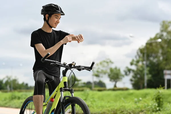 Smiling young Asian man cyclist resting after riding bicycle and checking sport activity progress data on smartwatch.