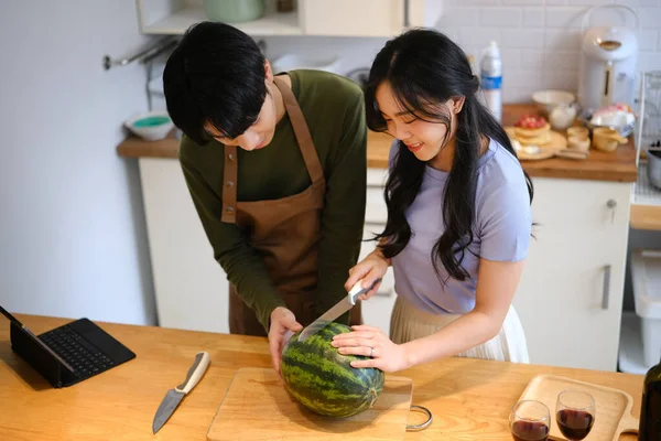 笑顔のアジア人男性は ガールフレンドが自宅のキッチンでスイカを切るのを手伝っています 関係性と楽しい時間を共にコンセプト — ストック写真