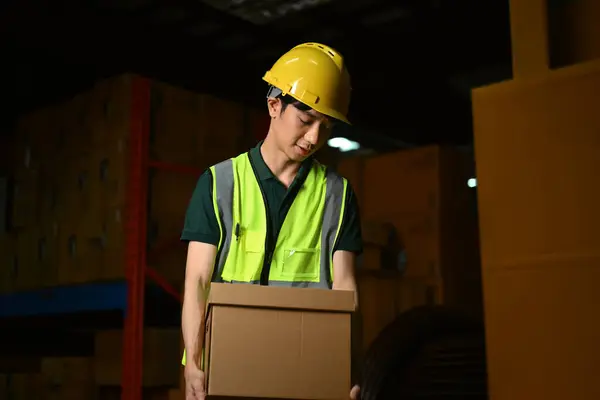 Warehouse worker holding cardboard box walking through retail warehouse full of packed boxes and goods.