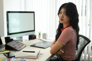 Grafik stüdyosunda boş bilgisayar monitörünün önünde oturan ve kameraya gülümseyen genç tasarımcının portresi..