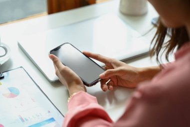 Yakın çekim görüntüsü, genç bir kadını ofis masasında otururken ve akıllı telefon kullanırken çekmiş..