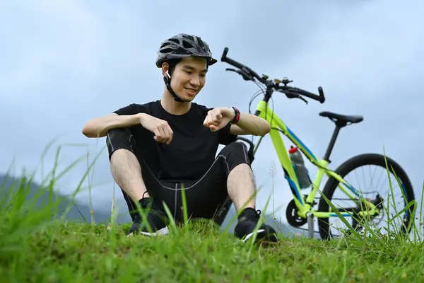 Satisfied man cyclist taking break from riding bicycle and checking sport activity progress data on smartwatch.