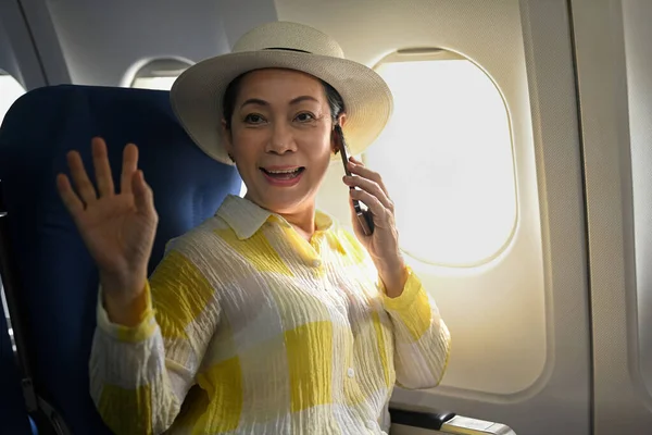 Positive middle aged woman tourist passenger talking on mobile phone and waving hand to camera.