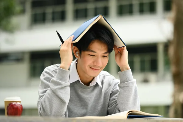 ハッピーアジアの学生は キャンパスで試験のために彼の頭に本を置く男 ライフスタイル — ストック写真