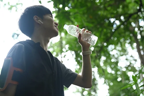 Happy asian male runner drinking water from a bottle, resting after trail running.