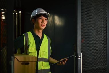 Endüstriyel fabrikada dikilirken çelik yelek ve karton kutu taşıyan yakışıklı bir erkek işçi..