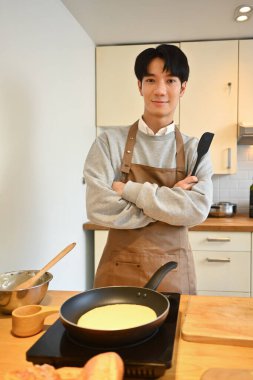 Elinde spatulayla önlük giyen yakışıklı Asyalı adamın portresi modern mutfakta kahvaltı hazırlıyor..