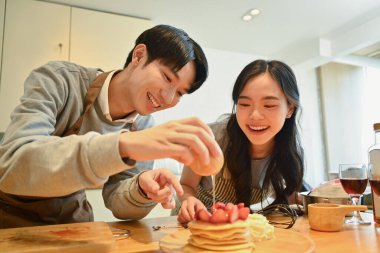 Neşeli genç çift lezzetli kreplere bal döküyor ahşap mutfak masasında çilek ile.