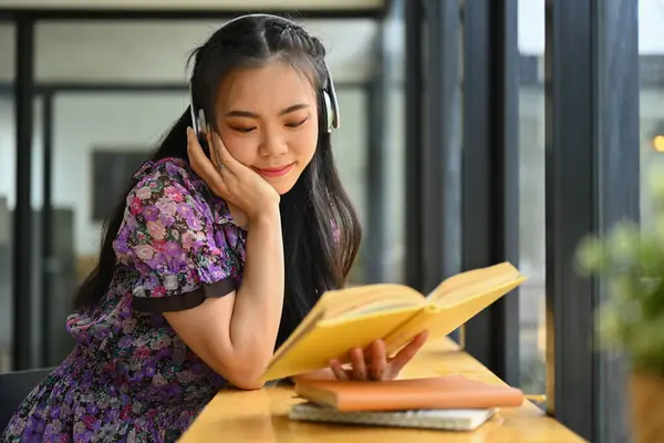 Pretty young woman college student listening to music in headphones and reading book in coffee shop.