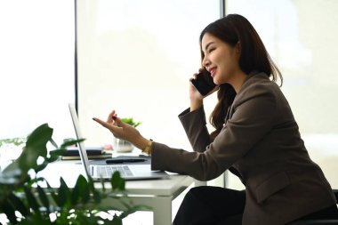 Güzel kadın yatırımcı danışmanı laptop ekranına bakıyor ve iş yerindeki müşteriyle cep telefonundan konuşuyor..