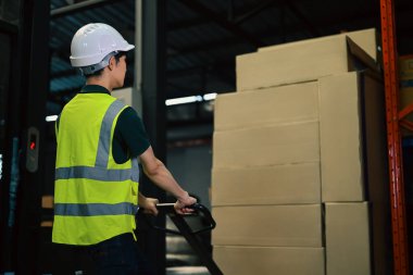 Depoda güvenlik kasketi takmış bir adam bir palet kamyonunu eşya dolu raflardan geçiriyor..