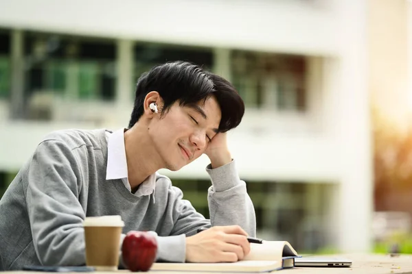 Carefree male university student listening to music on his earphones with closed eyes during reading book outdoor.