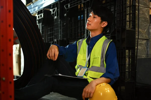 Tired man warehouse worker in jacket leaning against shelve while having short break.