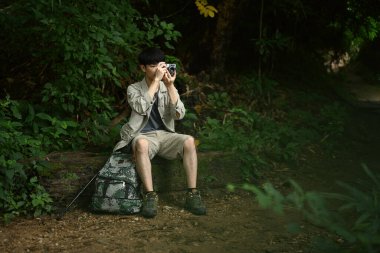 Erkek gezgin ağaç gövdesinde oturuyor ve kamerayla doğa manzarasını çekiyor. Seyahat ve macera konsepti..