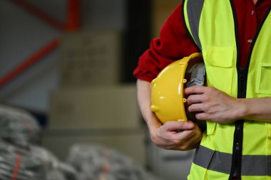 Güvenlik yeleği giyen tanınmayan bir depo çalışanı karton kutu yığınının yanında duruyor..