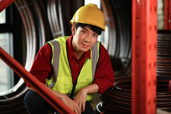 Asian male manager wearing hardhat and reflective jacket sitting in a warehouse and smiling to camera.