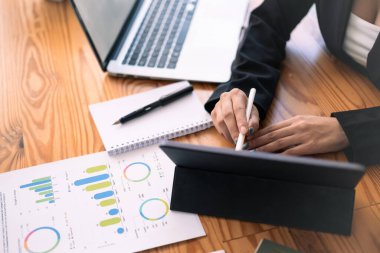 İş kadını istatistik belgelerini analiz ediyor ve ahşap ofis masasında dijital tablet kullanıyor.