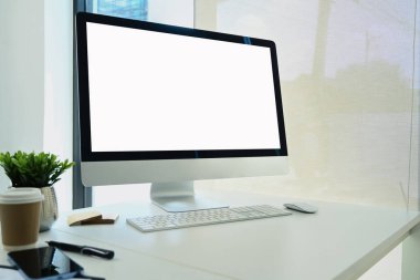 Boş ekranlı bilgisayar monitörü, kağıt bardak, akıllı telefon ve beyaz masa üzerinde saksı bitkisi olan rahat bir iş yeri..