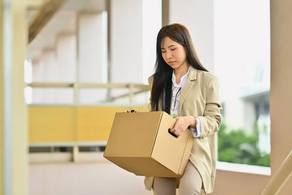 Upset female office worker leaving office with cardboard box full of belongings. Job loss, unemployed concept.