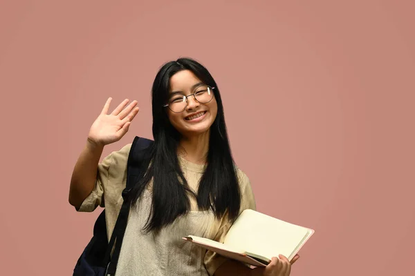 カメラで本と手を振るバックパックを持つ陽気な女子高生 そして学校のコンセプトに戻る — ストック写真