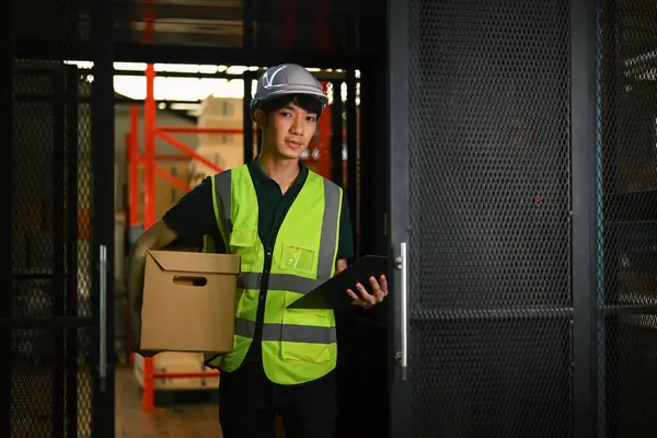 Asian male worker in hardhat and vest holding cardboard box while standing in industrial plant.
