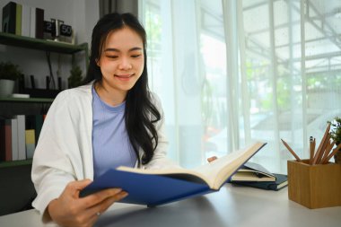 Günlük giysiler içinde, oturma odasındaki masada kitap okuyan mutlu genç Asyalı kadın. Eğitim ve eğlence etkinliği kavramı.