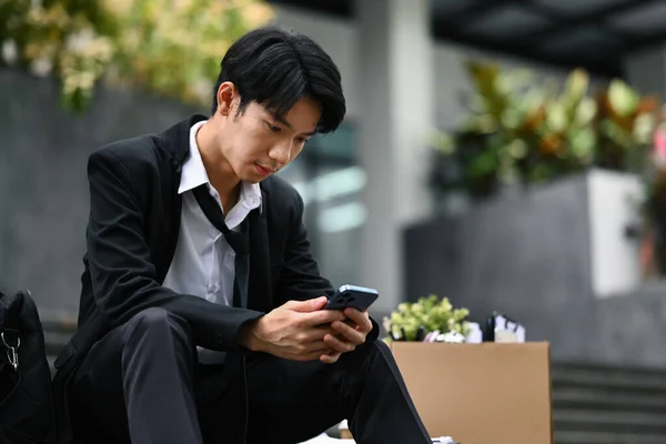 Upset asian male office worker sitting with box of personal stuff on stairs of office center and using phone. Unemployment concept.