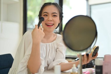 Neşeli genç bir kadın aynaya bakıyor ve yanaklarını fırçalayarak makyaj yapıyor. Güzellik, kozmetik ve yaşam tarzı konsepti.