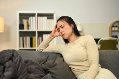 Mutsuz bir kadın kanepeye oturmuş, gözleri kapalı ve başına dokunuyor, şiddetli baş ağrısı ya da migrenden muzdarip..