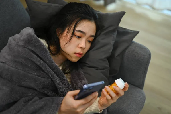 薬瓶を手に持っている病気の若い女性とインターネットでの治療に関する情報を検索するためにスマートフォンを使用 — ストック写真