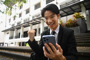 Heyecanlı genç işadamı akıllı telefona bakıyor, kazanan evet hareketi yapıyor, zafer ve başarıyı kutluyor..