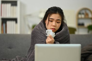 Kanepede battaniyeyle kaplanmış hasta genç bir kadın dizüstü bilgisayarında doktorla online istişare yapıyor. Teletıp kavramı.