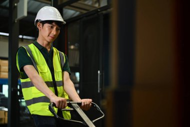 Güvenlik kasketli bir işçi, perakende ambarında satılık mallarla rafların arasından bir palet kamyonu çekiyor..