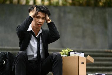 Erkek ofis çalışanı, elinde kişisel eşyalarla umutsuzca binanın merdivenlerinde oturuyor. İşsiz, depresif konsept.