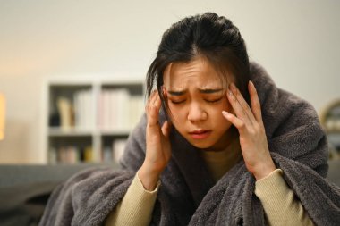 Kanepede battaniyeyle oturan mutsuz kadın şiddetli baş ağrısı ya da migrenden muzdarip..