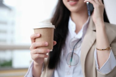 Tek kullanımlık kahve fincanıyla modern ofis binasının önünde cep telefonuyla konuşan bir iş kadını..